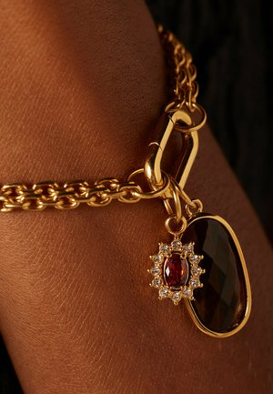 Gouden armband met een grote donkere edelsteen en een kleinere rode edelsteen omgeven door heldere stenen, die ingewikkelde details en texturen benadrukt.