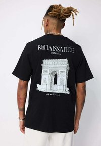 Homme avec des dreadlocks portant un t-shirt noir affichant un croquis blanc de l'Arc de Triomphe et les mots "Renaissance" et "Potentiel" dans le dos.