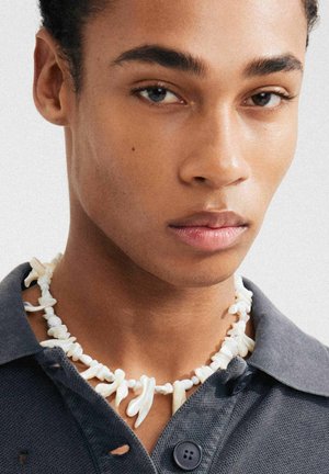 Halsband med texturerade vita element och små pärlor, bärs över en mörk skjorta med krage. Har oregelbundna former och naturliga material.