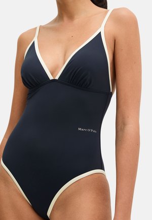 Frau trägt einen marineblauen Einteiler mit weißer Umrandung und dünnen Trägern, seitlich mit "Marc O'Polo" gebrandet.