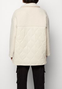 Off-white, fleecefodrad jacka med en quiltad beige nedre sektion. Har krage och långa ärmar. Dekorativ quiltmönster på botten.