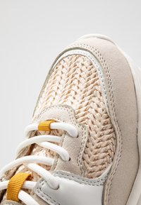 Sneakers med en vävd beige ovandel, ljusgrå mockadetaljer, vita snören och en gul etikett. Strukturerade mönster genom hela designen.