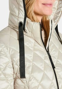 Gepolsterte, hellbeige Steppjacke mit hohem Kragen, schwarzen Reißverschlüssen und kontrastierenden schwarzen Kordelzügen. Glatte Textur mit glänzendem Finish.