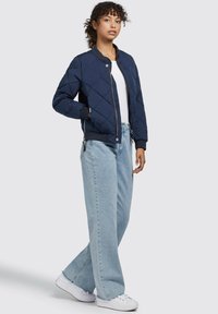 Chaqueta bomber acolchada en navy con cremallera frontal, puños y banda elástica, combinada con jeans de pierna ancha en azul claro y zapatillas blancas.