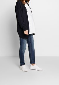 Marinblå stickad cardigan med öppen front, långa ärmar och ribbade kanter, tillsammans med en vit t-shirt, blå jeans och vita sneakers.