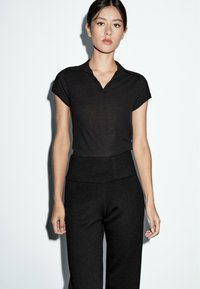 Jonge vrouw met donker haar dat naar achteren is vastgebonden, in een zwart kortmouw collared top en hoog gevestigde zwarte broek, staande tegen een witte achtergrond.