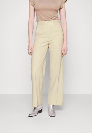 Pantalons larges beiges en tissu lisse, dotés d'une taille haute et de plis marqués, associés à des bottines argentées brillantes.