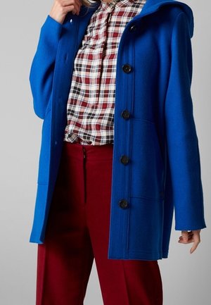 Vrouw draagt een blauwe jas met knopen over een rood-wit geruit shirt en rode broek, houdt met één hand de kraag van de jas vast.