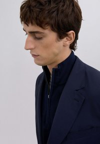 Blazer navy sobre un suéter de punto con cuello alto y cremallera. Material suave y estructurado; textura sutil en el suéter; diseño ajustado.