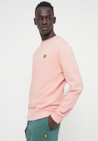 Φούτερ ανοιχτό ροζ με λαιμόκοψη crew, με μικρό κεντητό κίτρινο λογότυπο στο στήθος. Το ύφασμα φαίνεται απαλό και με υφή.