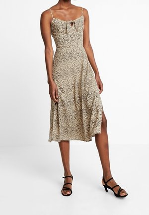 Woman wearing a sleeveless beige and black leopard print midi dress with a front tie detail and black strappy heeled sandals.