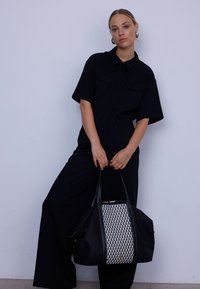 Čierna taška s textúrovaným strieborným tkaným panelom, vybavená dvojitými rukoväťami a zipsovým uzáverom. Modelka má na sebe čierny overal s golierom.