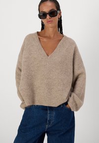 Pulover bej, tricotat cu model cu caneluri, cu decolteu adânc în V și croială supradimensionată, asortat cu blugi casual de culoare albastru închis. Însoțit de ochelari negri de soare.