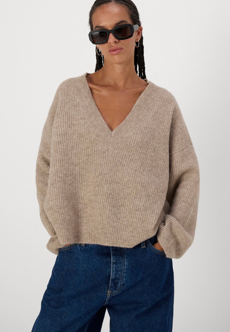 Beige, geripptes Strickpullover mit tiefem V-Ausschnitt und oversized Schnitt, kombiniert mit lässigen dunkelblauen Jeans. Begleitet von schwarzen Sonnenbrillen.