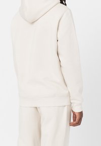 Krémově zbarvená mikina s kapucí, měkkou texturou, dlouhými rukávy a stahovací šňůrkou u kapuce. Jednoduchý design s gumovanými manžetami.