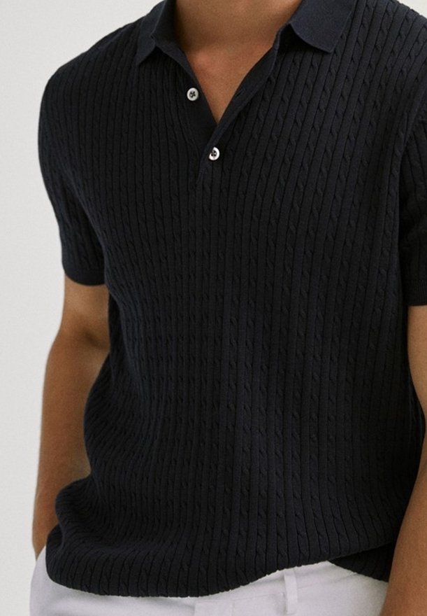 Homme portant un polo en maille texturée noire à manches courtes avec des boutons blancs, associé à un pantalon blanc.