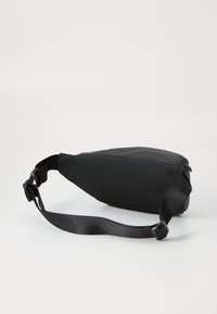 Sac bandoulière noir vu de côté, montrant la sangle réglable et la fermeture éclair. Le sac a un design épuré et minimaliste.