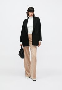 Blazer negro extragrande, cuello alto blanco, pantalones de pana beige y bolso negro. El calzado es blanco con detalles en negro. Diseño sencillo y moderno.