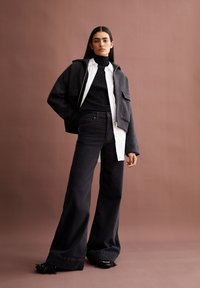 Grå kort jacka över en svart stickad polotröja och vit skjorta med knappar, i kombination med vida svarta byxor och klackskor med tofsar.