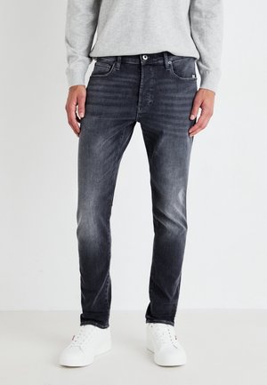 Homme portant un jean slim noir délavé, un pull gris clair et des baskets blanches, debout devant un fond clair uni.