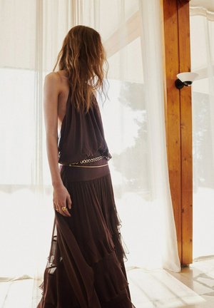 Femme aux longs cheveux portant une robe marron sans manches superposée avec une ceinture dorée, debout à l'intérieur près de rideaux légers et de poutres en bois.