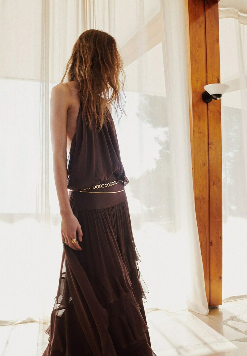 Femme aux longs cheveux portant une robe marron sans manches superposée avec une ceinture dorée, debout à l'intérieur près de rideaux légers et de poutres en bois.