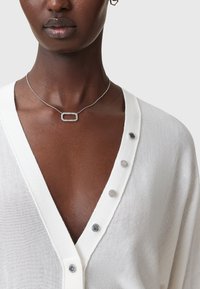 Valkoinen, kevyt neuletakki, jossa on syvä V-pääntie, seitsemän hopeista nappikoristetta, mustan ihon sävyn päällä, ja siihen kuuluu herkkä kaulaketju.