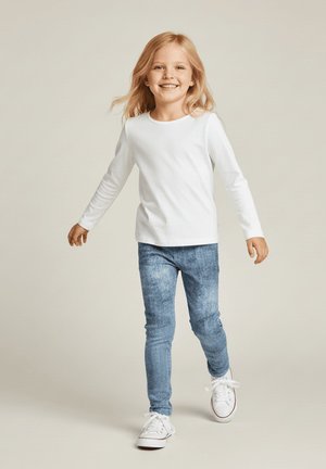 Małe dziecko z długimi blond włosami spacerujące, ubrane w białą koszulę z długim rękawem, niebieskie dżinsy i białe trampki na neutralnym tle.