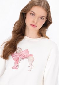 Fehér pulóver, amelyen egy nagy rózsaszín masnit viselő kutya nyomtatott mintája látható. Az anyag puhaságnak tűnik, laza szabású, kerek nyakkivágással.