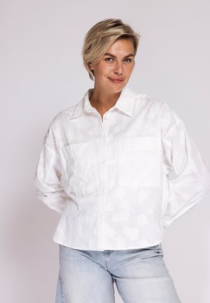 Frau mit kurzen blonden Haaren, die ein strukturiertes weißes Hemd mit Knöpfen und hellblaue Jeans trägt, steht mit den Händen hinter dem Rücken.