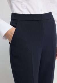 Námořnické modré kalhoty na míru s hladkou texturou, plochým předním dílem a bočními kapsami, doplněné bílou košilí s knoflíky.