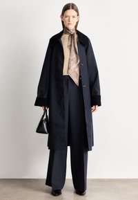 Tengerészkék hosszú kabát fekete gallérral, világosbarna felsővel és mintás sállal. Széles szárú tengerészkék nadrág és egy kis fekete kézitáska teszi teljessé az öltözéket.