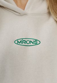 Kremowy hoodie z zielonym haftowanym logo "MIRÓNS" w owalnym kształcie, wykonany z miękkiego materiału o teksturowanym wykończeniu.