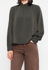 Blouse vert foncé à manches longues avec un col mao et des poignets boutonnés ; présente une coupe ample et une texture douce. Portée avec un pantalon marron.