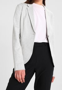 Grå kavaj med skärning, enkel knappstängning och slagkrage, bärs över en ljusrosa skjorta och svarta byxor.