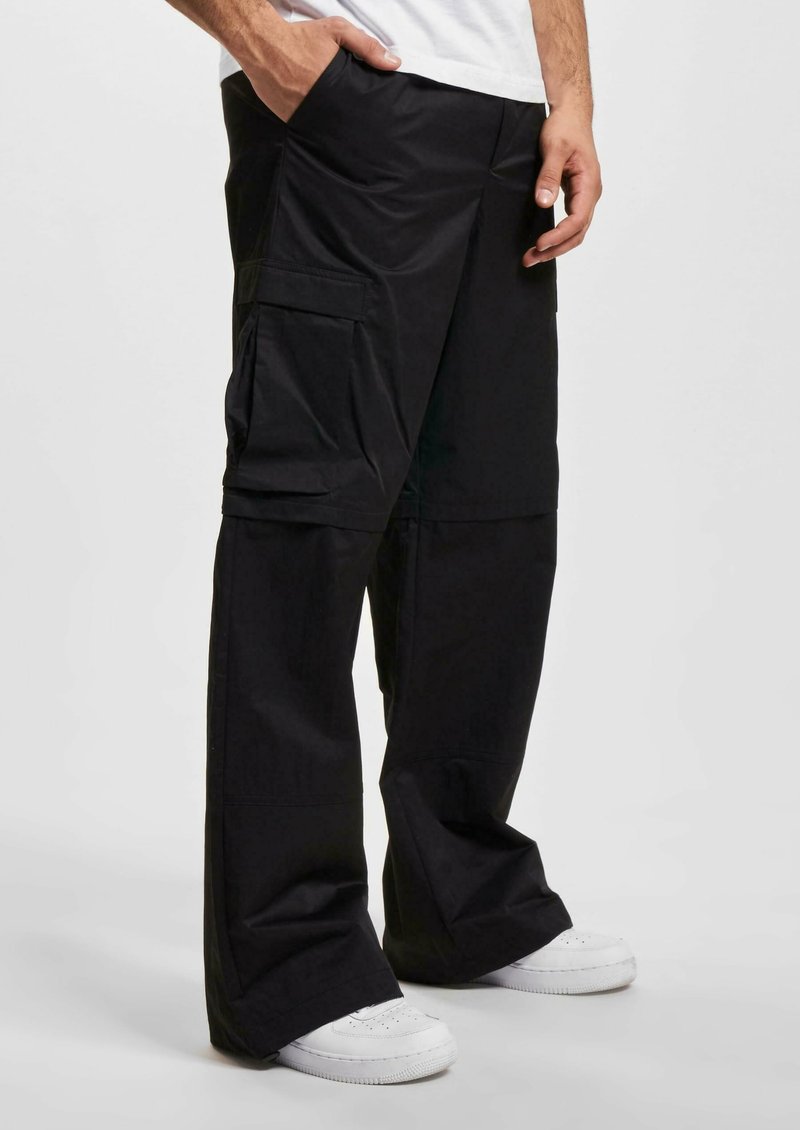 Pantalons cargo noirs à jambes larges, dotés de plusieurs poches et d'un tissu lisse et durable. Portés avec des baskets blanches.