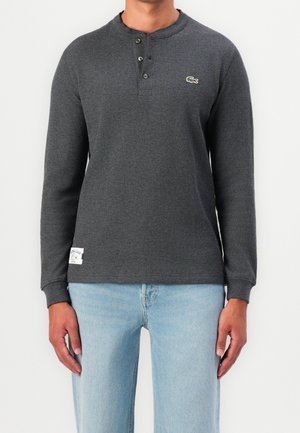 Mann trägt ein dunkelgraues Langarm-Henley-Shirt mit drei Knöpfen und hellblaue Jeans, steht vor einem schlichten weißen Hintergrund.
