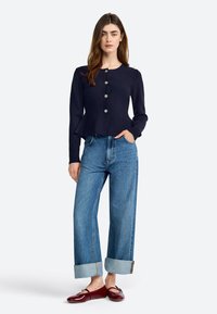 Navy long-sleeve cardigan with silver buttons, peplum design, paired with loose blue denim jeans featuring cuffed hems and burgundy flats.