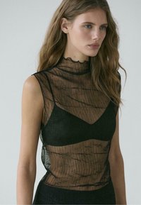 Černý průsvitný top s vysokým, zvlněným výstřihem a tenkou látkou. Má vrstvený design přes černou podprsenku. Texturovaný, lehký materiál.