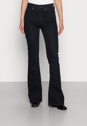 Femme portant un jean noir évasé taille haute avec poches avant et poche monnaie, associée à des chaussures noires sur un sol gris et un fond blanc.