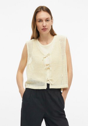 Vrouw met lang haar draagt een crème kleurig gebreid mouwloos vest over een licht overhemd en zwarte broek, staand met handen in de zakken tegen een witte achtergrond.