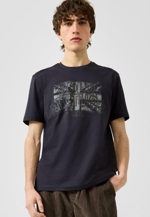Junger Mann trägt ein schwarzes T-Shirt mit verblasstem Text "BOLD ATTITUDE" und einem grafischen Design, kombiniert mit braunen Cordhosen.