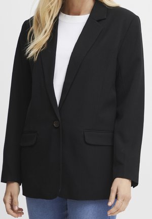 Femme portant un blazer noir à un seul bouton sur une chemise blanche et un jean bleu, montrée du cou aux hanches sur un fond uni.