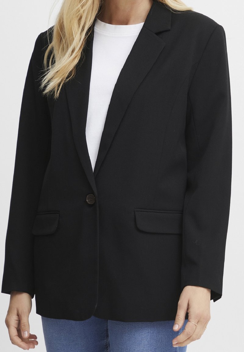 Femme portant un blazer noir à un seul bouton sur une chemise blanche et un jean bleu, montrée du cou aux hanches sur un fond uni.