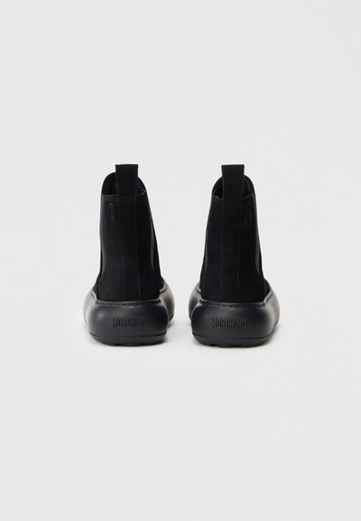 Crne nubuk čizme do gležnja s elastičnim bočnim panelima i okruglim gumenim potplatima. Karakteristike uključuju otisnutu oznaku na peti, minimalistički dizajn i glatku teksturu.