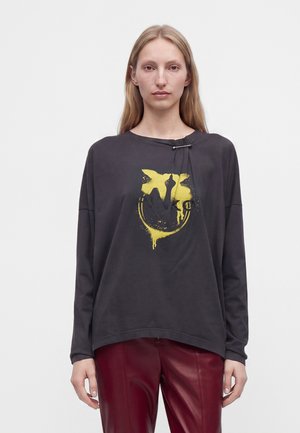 Femme portant une chemise noire ample à manches longues avec un imprimé abstrait jaune et un pantalon en cuir rouge, debout devant un fond blanc uni.