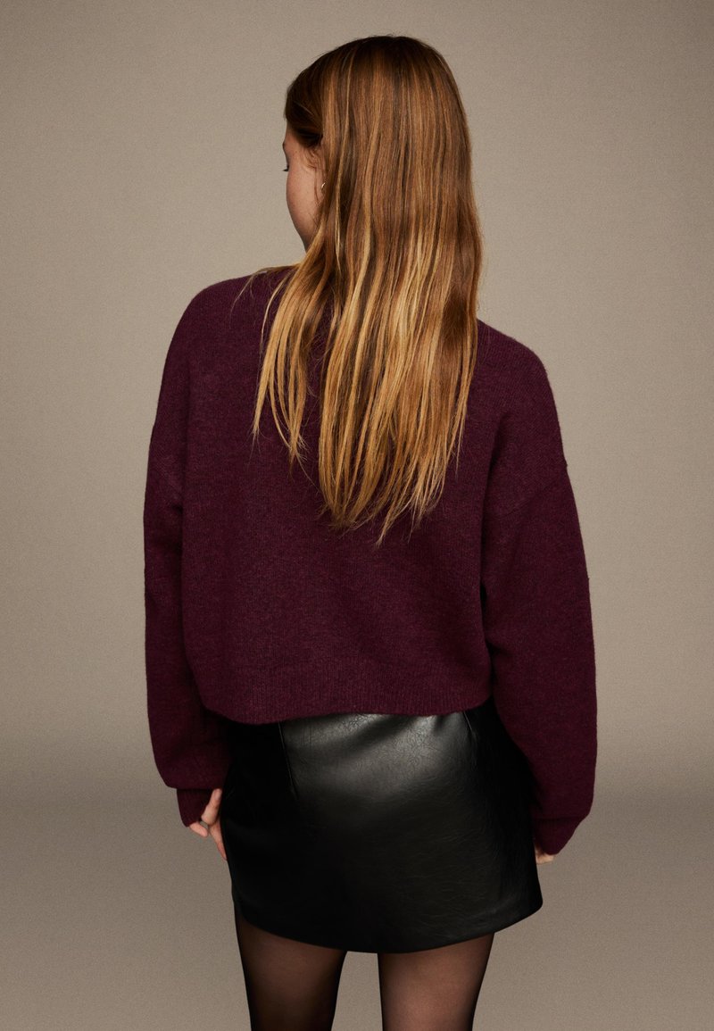 Bordeauxfarbener oversized Strickpullover mit weiten Ärmeln, kombiniert mit einem schwarzen Leder-Minirock und transparenten schwarzen Strumpfhosen.