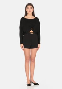 Top nero in maglia con design frontale attorcigliato, abbinato a pantaloni corti neri a vita alta con dettaglio a fibbia dorata. Aspetto elegante e aderente.