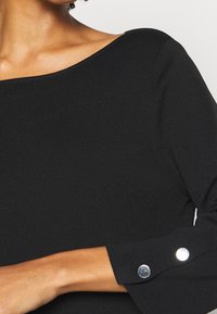 Haut noir côtelé avec un large col et des manches longues. Présente des accents de boutons en argent aux poignets. Le tissu semble doux et texturé.