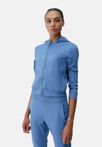 Ljusblå zip-up huvtröja med huva, figurnära design, ribbade muddarna och sömmar som skapar ett texturerat mönster. Slätt, stretchigt tyg.